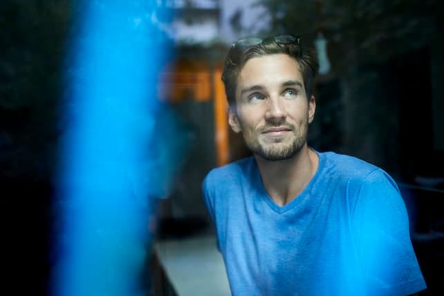 a portrait of a man in a blue t-shirt sitting in a dark room and thinking